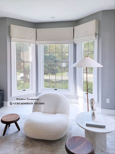 Bright and inviting room in Closter, NJ, showcasing a bay window adorned with neutral-colored roman shades. The space includes a plush white chair, a small round table with a lamp, and a modern, minimalist design.