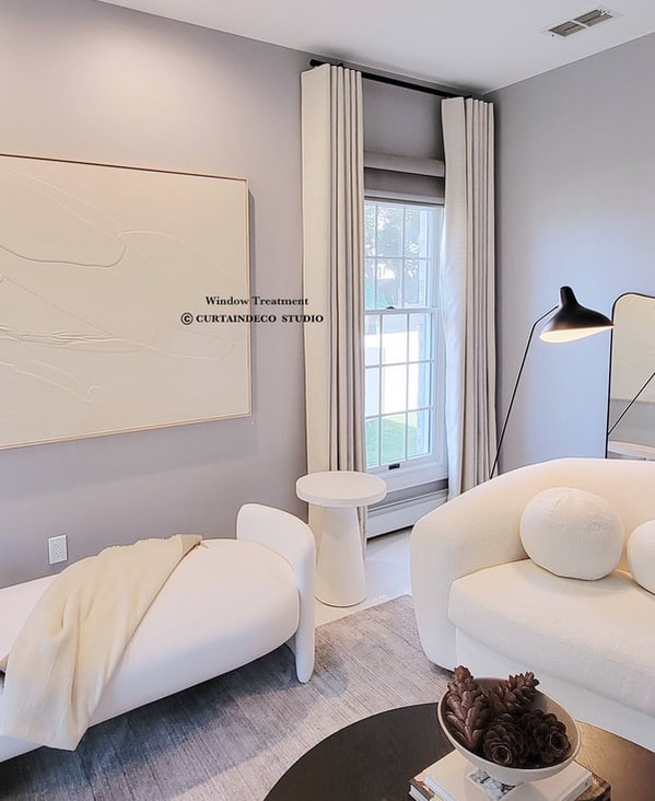 Modern and cozy room in Closter, NJ, featuring elegant floor-to-ceiling white curtains framing a tall window with a gray roman shade. The room is complemented by a stylish white lounge chair, a round side table, and minimalist decor.