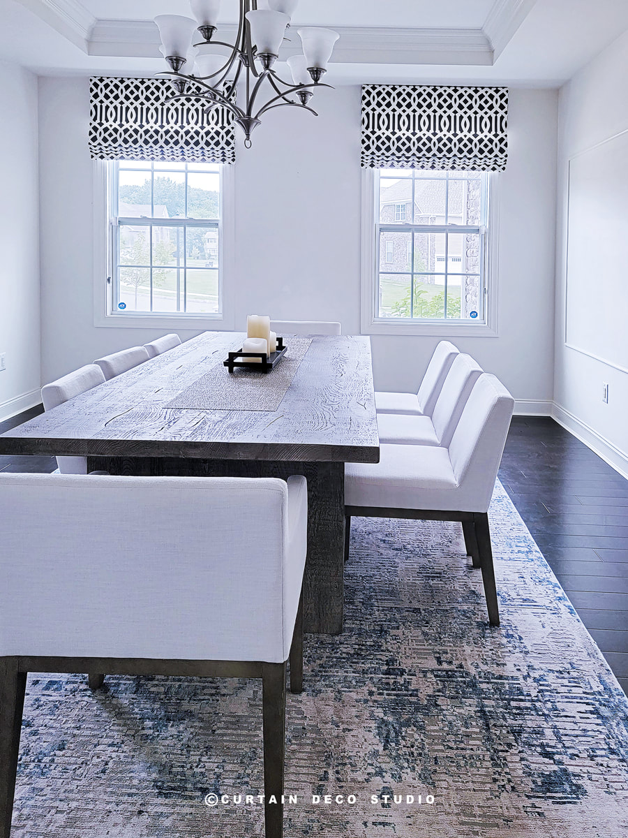 A dining room with a large wooden table and white cushioned chairs. The windows are adorned with black and white geometric fabric Roman shades, enhancing the room's modern and elegant aesthetic.
