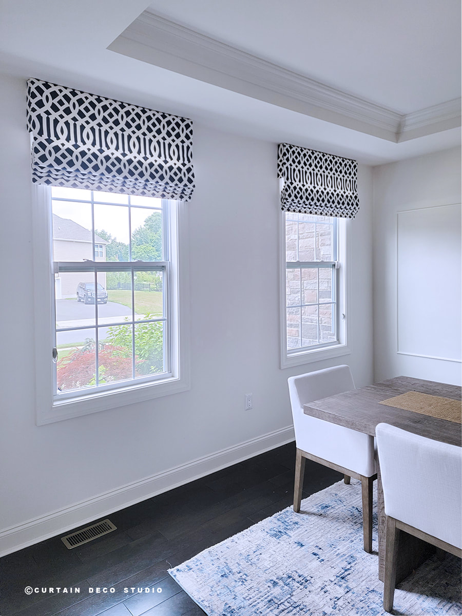 Close-up view of a dining room window with a black and white geometric fabric Roman shade. The window provides a clear view of the outdoor scenery, while the Roman shade adds a touch of sophistication and style to the room.